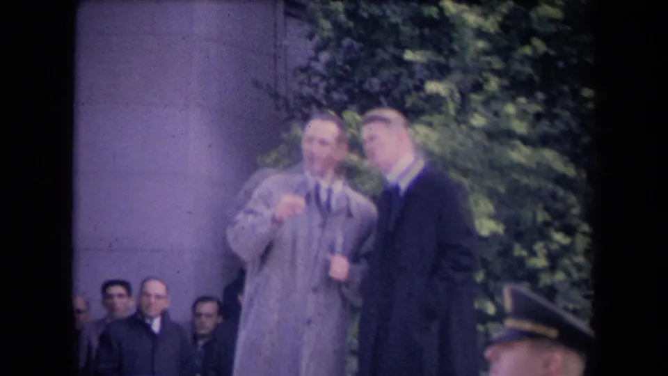 Two men in formal coats stand near a stone structure with onlookers behind them, one appearing to gesture during the outdoor event.