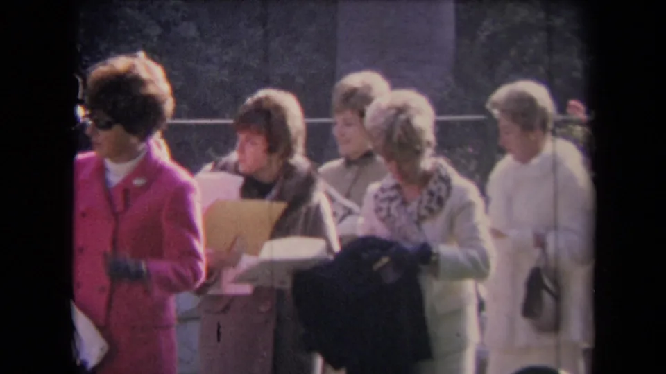 Women in 1960s formal clothing, including a bright pink suit and fur-trimmed coats, hold papers while gathered outdoors.