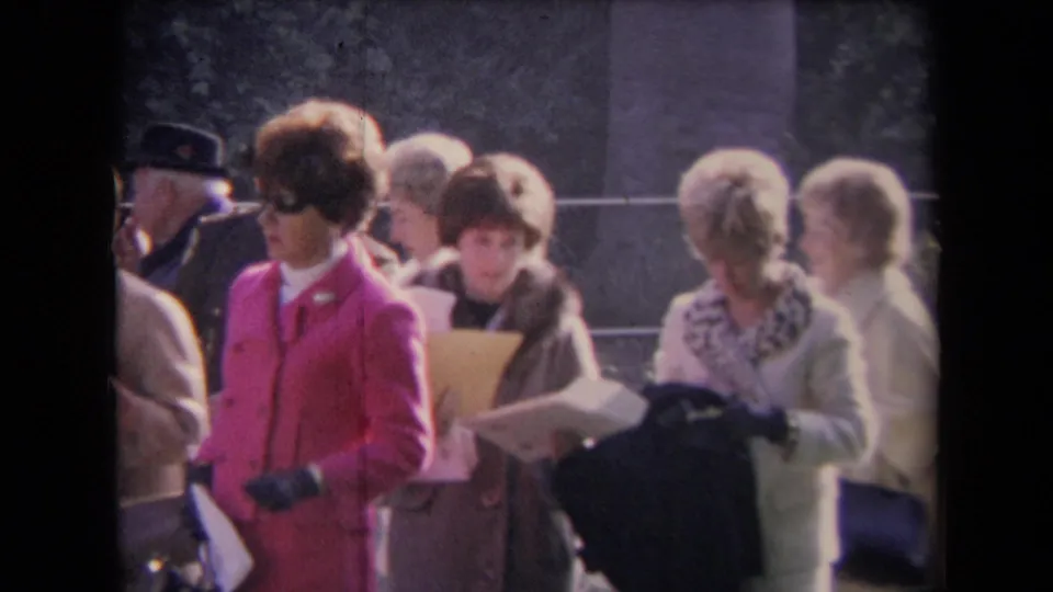 Outdoor 1960s home-movie frame showing women in formal coats, one in a vivid pink suit and sunglasses, with papers and railings behind them.