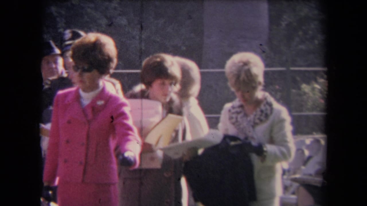 A woman in a pink suit stands near women holding papers, with an outdoor railing and trees behind them.