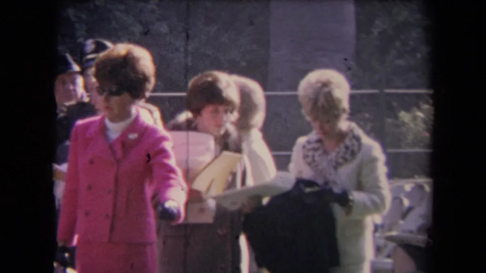 A woman in a bright pink suit and sunglasses stands among women handling papers at an outdoor civic gathering in 1960s footage.