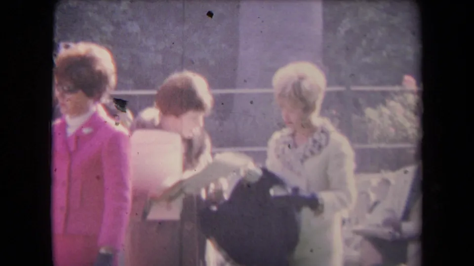 Women in pale coats look down at papers and a dark bag outdoors, while the pink-suited figure is partly visible at frame left.