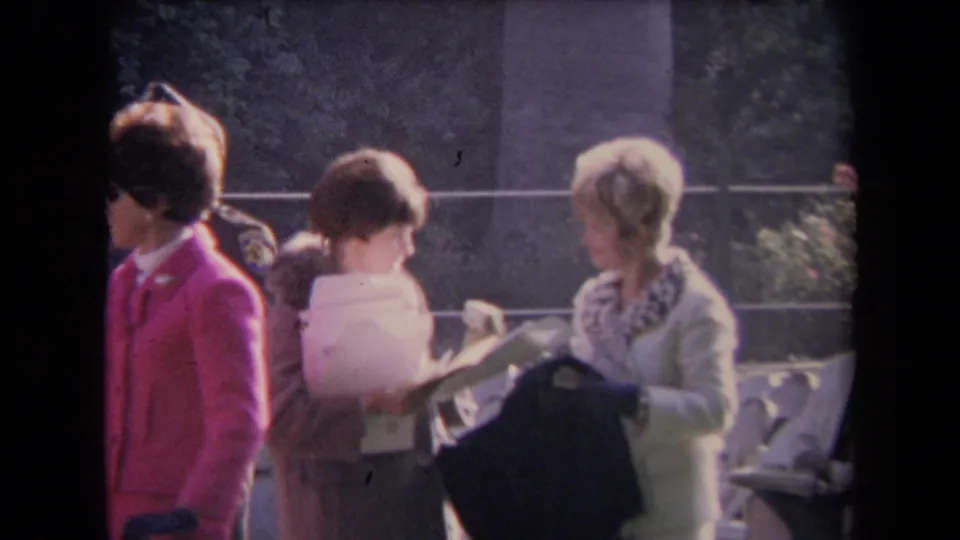 A group of women in 1960s coats cluster near papers and a dark bag, with railings and trees in the background.