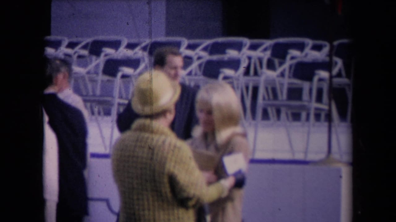 A woman in a yellow hat and plaid coat embraces a blonde woman near rows of empty folding chairs at a public event.