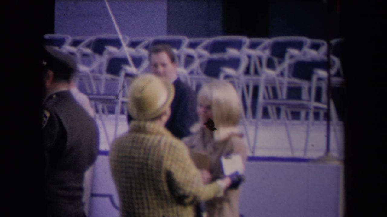 The greeting continues as a uniformed figure passes at left and rows of empty folding chairs fill the background.