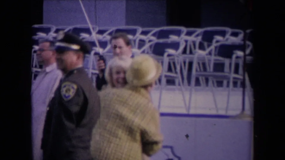 Uniformed personnel and other attendees move through the foreground as the woman in the yellow hat turns near the chair rows.
