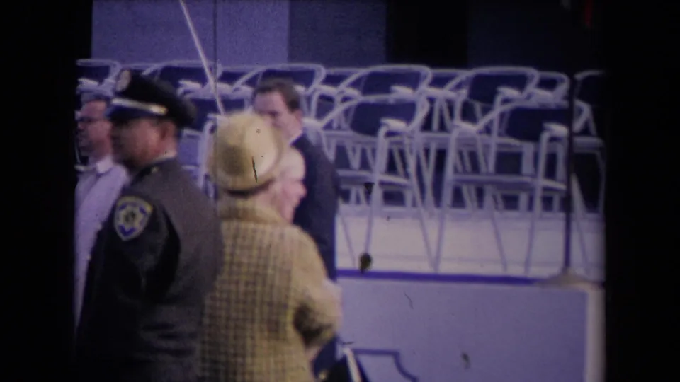 1967 home-movie frame of a uniformed officer near a woman in a plaid coat and hat at a large public event.