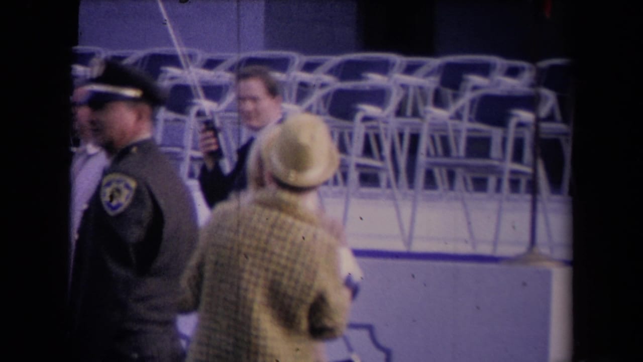 Home-movie view of a uniformed officer, a woman in a hat, and suited attendees near rows of stacked chairs.