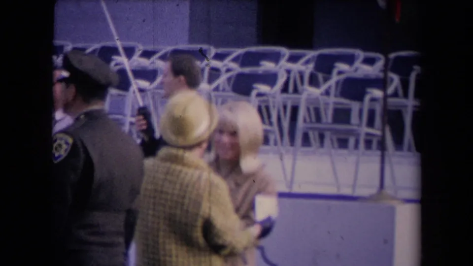 1967 event frame showing a uniformed officer, two women in formal clothing, and stacked chairs in the background.