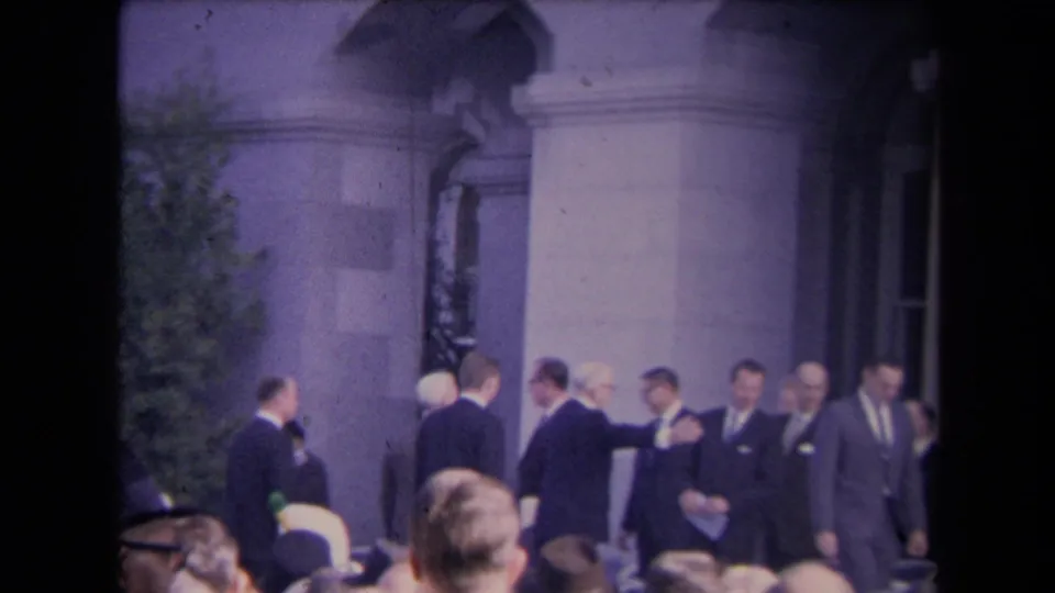 Wider home-movie frame of suited attendees and a woman in a hat near a columned entrance at a formal public event.