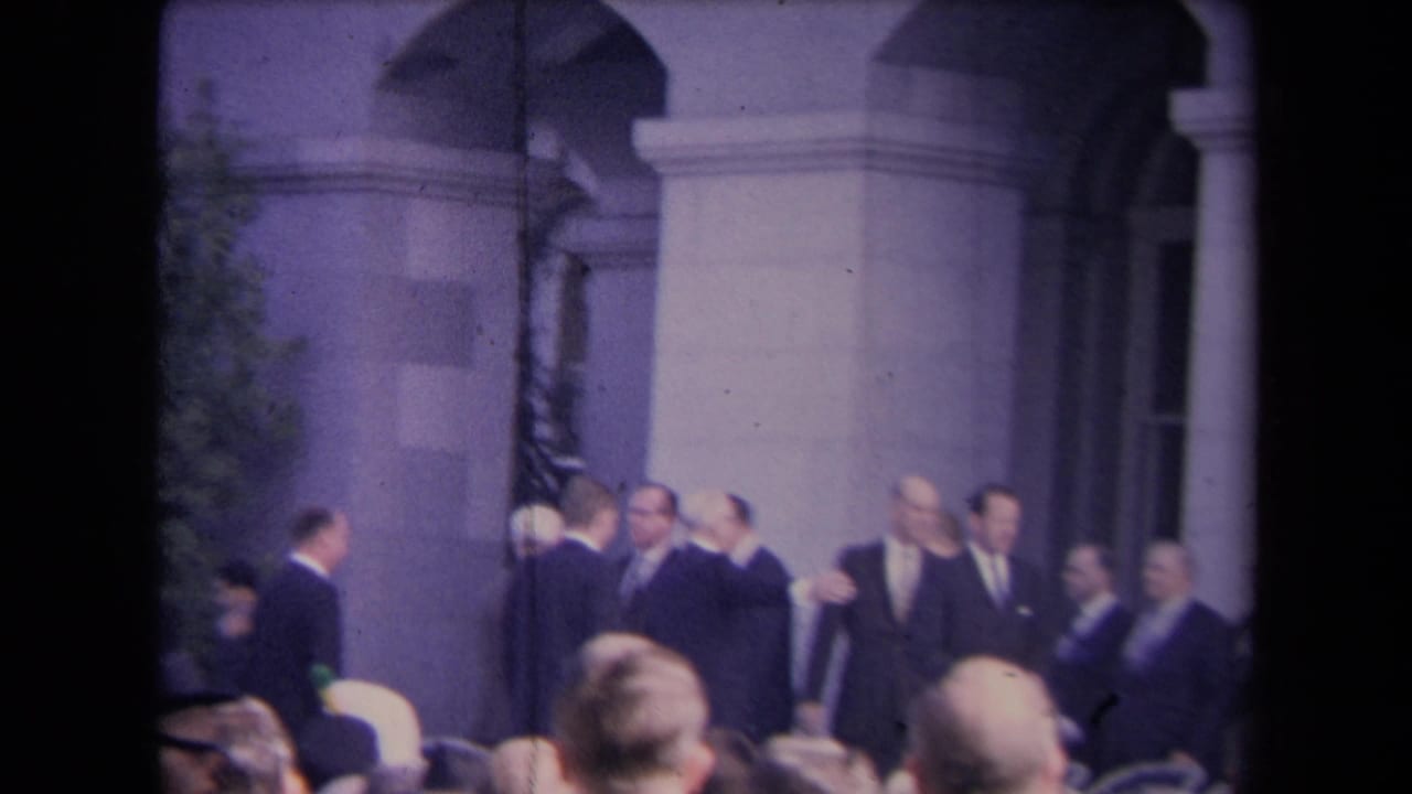 Men in dark suits greet each other before a columned arched building as a crowd watches.