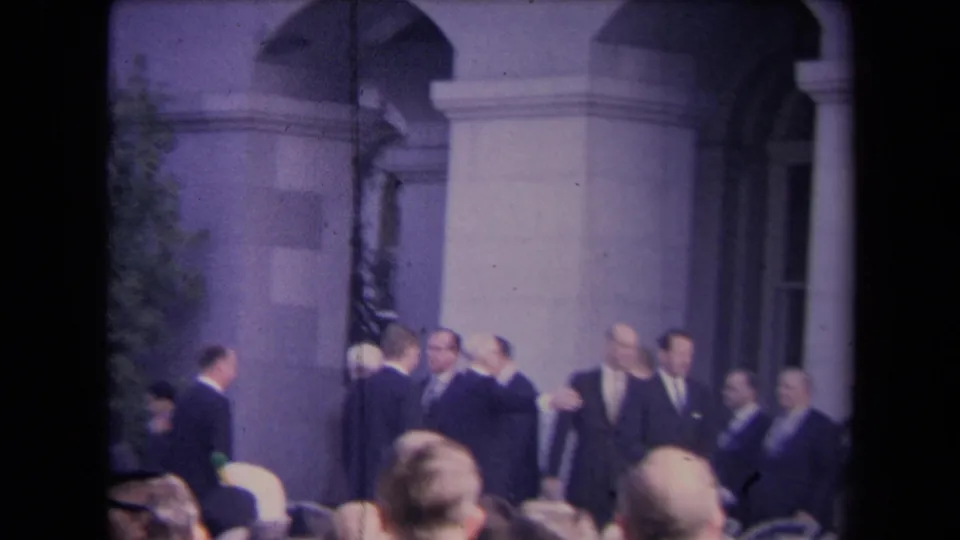 1967 8mm home-movie frame of suited men gathered outside a grand arched facade, with onlookers in the foreground.
