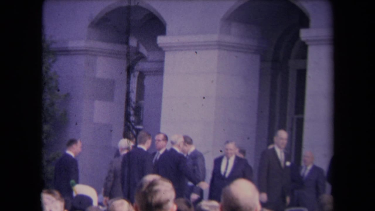A group of suited men stands in conversation outside a large arched building entrance.