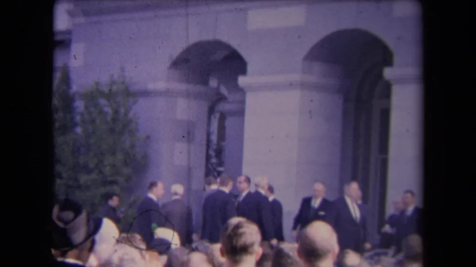 Handheld 8mm view of a formal group near a columned facade, with spectators' heads and hats visible in the foreground.