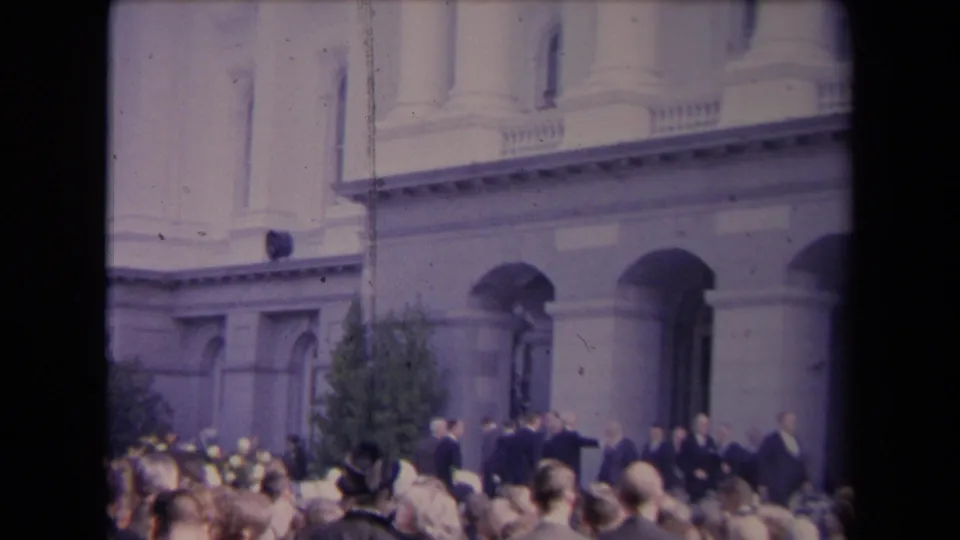 Wider 8mm frame showing spectators facing a small group of suited men near arched entrances and a columned facade.