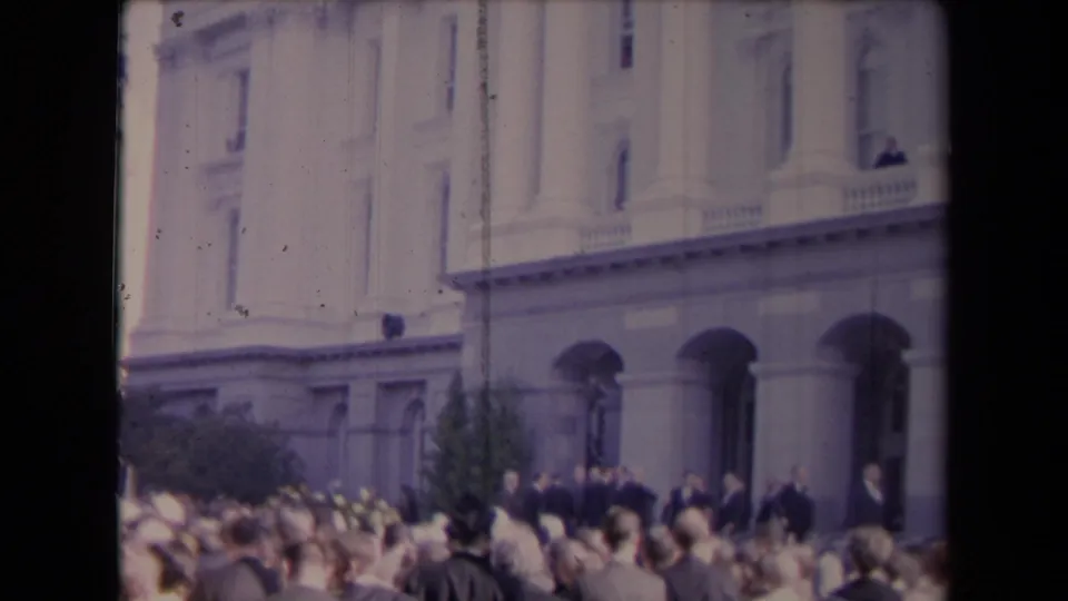 A dense public crowd stands before a grand classical building, with suited figures visible near the arched entryways.
