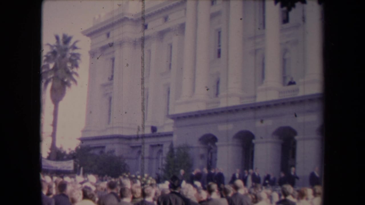Crowd in front of a columned building with a palm tree at the left.