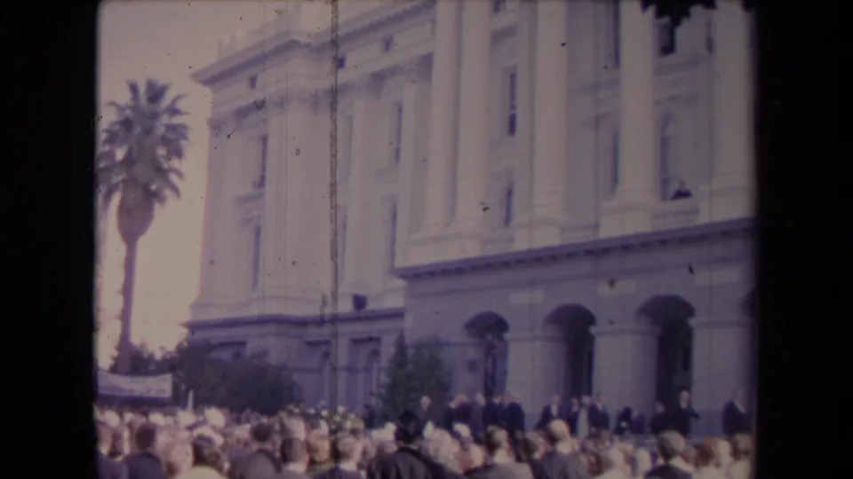 The crowd fills the foreground outside the building, with arches, columns, and a tall palm tree visible in the background.