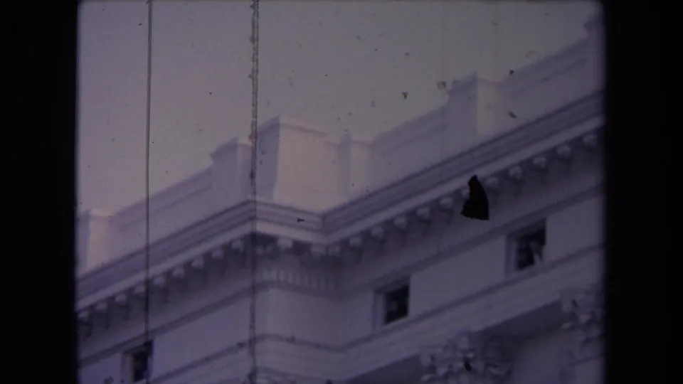 The camera tilts or frames upward on the building’s upper facade, showing windows, cornice detail, and visible film wear.