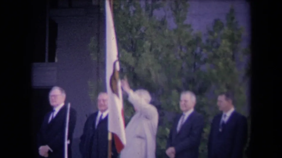 1967-era outdoor political ceremony frame with Ronald Reagan appearing to wave beside officials, a flagpole, and trees.