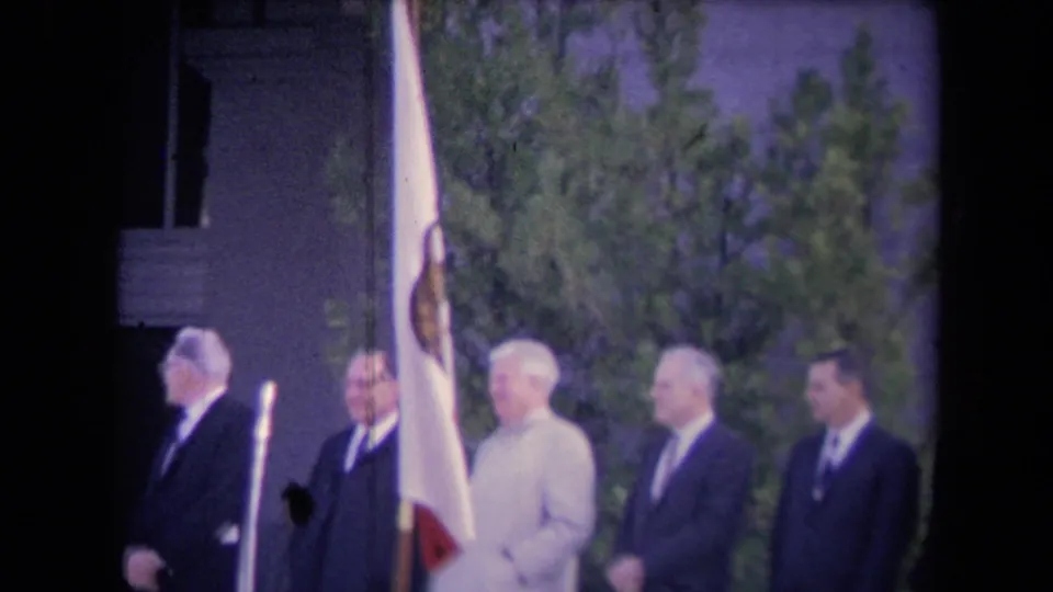 Outdoor 8mm ceremony view showing Ronald Reagan in a light suit among suited officials, with a flagpole and trees behind them.