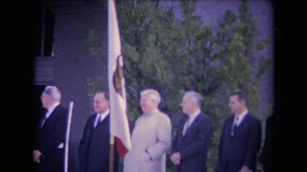 Line of suited men standing near a flag beside a columned building.