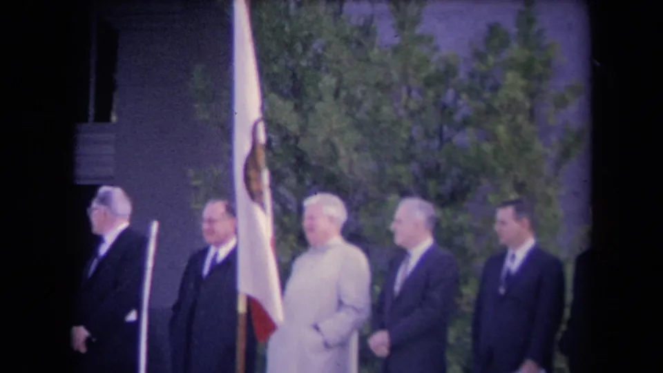 Formal outdoor ceremony with suited men, one light-coated figure, and a red-and-white flag near a columned public building.