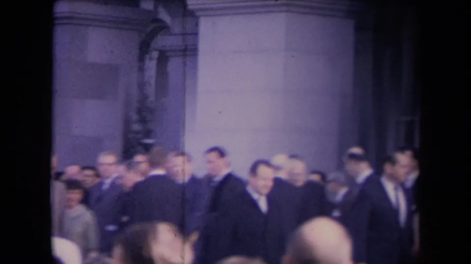 Suited attendees cluster near tall columns and a doorway, with several faces visible in soft-focus 8mm footage.