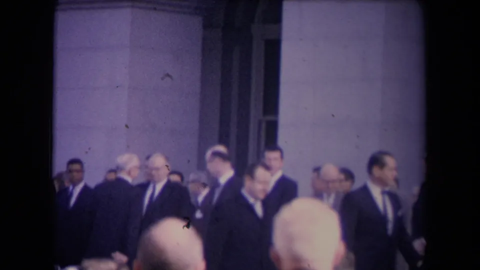 A handheld view from the crowd shows suited men gathered before stone columns, partly blocked by heads in the foreground.
