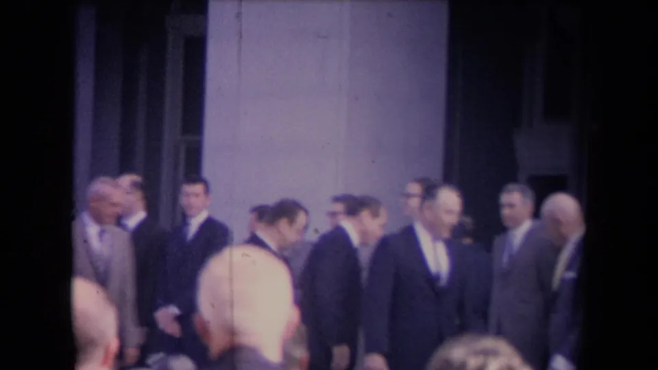 Men in dark suits stand and move near a columned building while viewers in the foreground watch the ceremony.