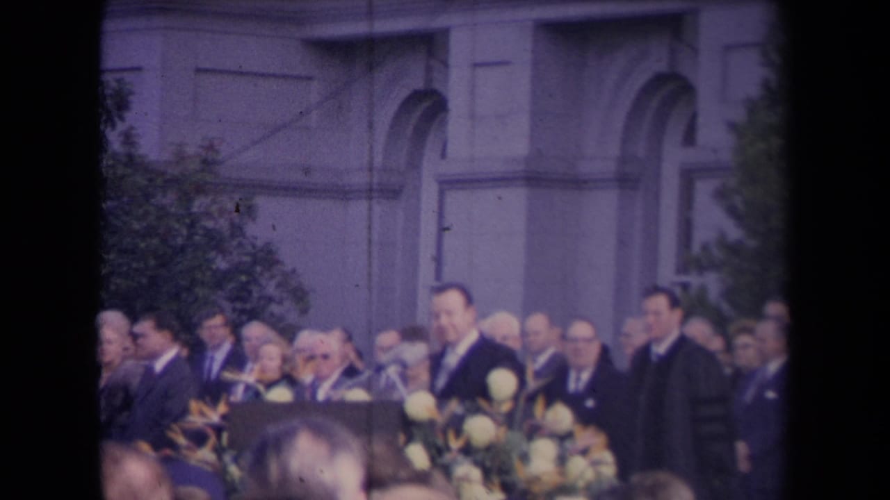Crowd of formally dressed men standing near flowers and arched architecture.