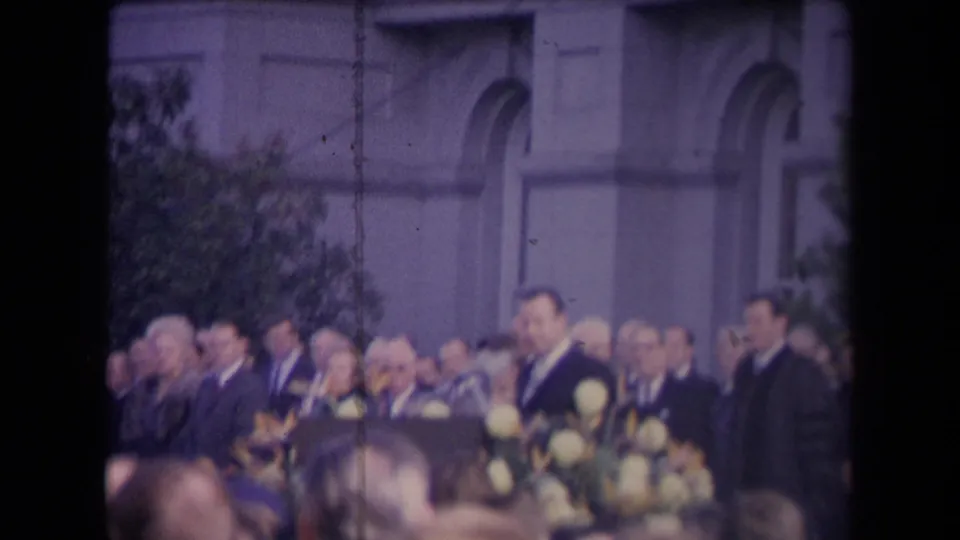 Home-movie frame from the supplied 1967 political event showing a crowd, suited officials, flowers, and classical-style architecture.