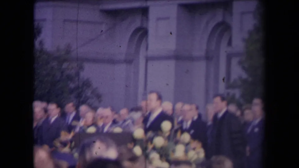 Soft, purple-tinted 8mm or Super 8 frame of a formal political crowd with flowers and arched building details behind them.