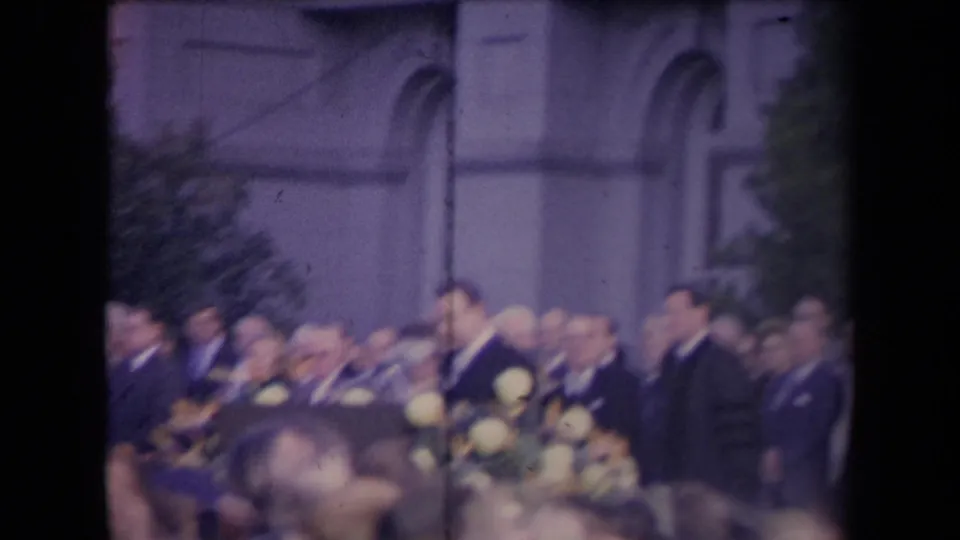 Soft 8mm frame of a formal outdoor crowd in front of arched architecture, with flowers and suited attendees visible.