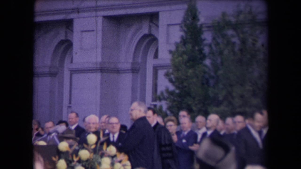Robed man and suited attendees stand near microphones and flowers outdoors.