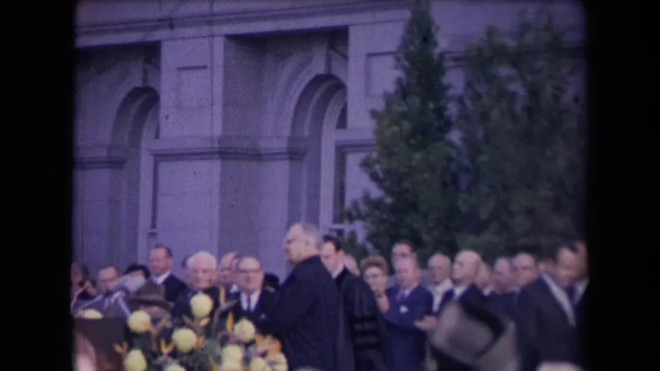 Formal ceremony scene, metadata-linked to Sacramento in 1967, with robed participant, microphones, flowers, and crowd before arched architecture.