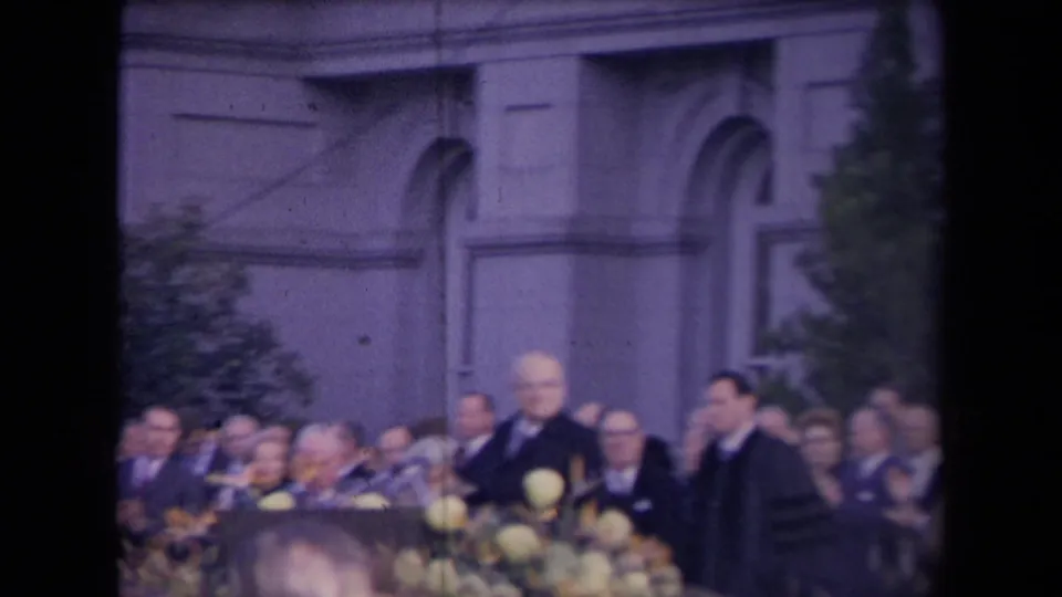 Soft-focus 8mm view of a formal public ceremony with audience, robed figure, microphones, flowers, and classical exterior details.