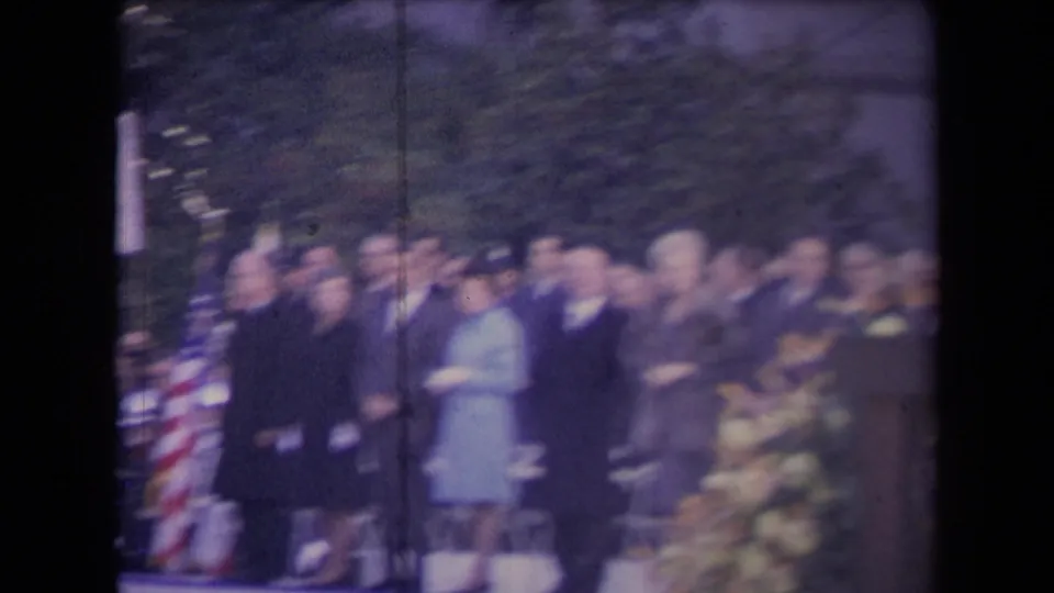 Blurred 8mm frame of a formal outdoor gathering with suited attendees, a woman in a light coat, an American flag, trees, and flowers.