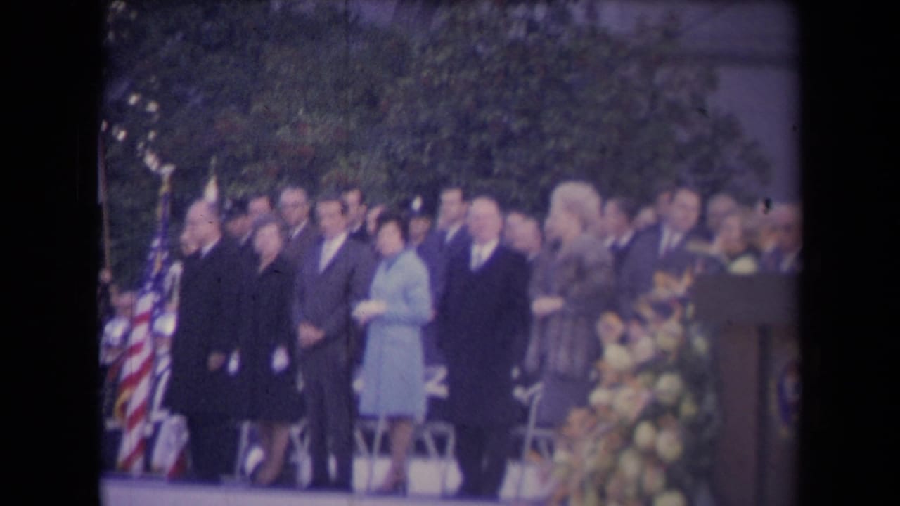 Formal crowd standing outdoors beside an American flag and flowers.
