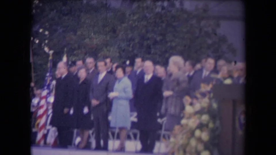1967 8mm frame of formally dressed adults standing at an outdoor civic ceremony with flags, trees, chairs, and flowers visible.