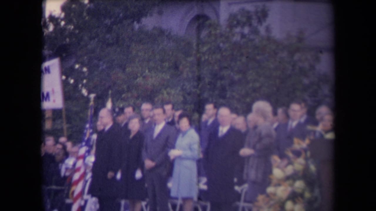 Formally dressed adults stand in a line at a 1960s outdoor ceremony, with American flags, a partial sign, trees, and flowers in view.