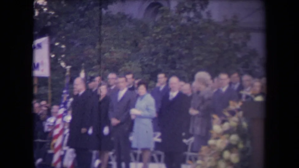 Home-movie frame showing a formal outdoor gathering, with suited men, a woman in a light coat, flags, trees, and chairs visible.