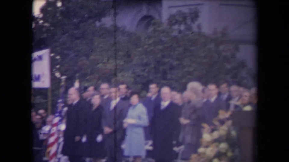 Soft 8mm frame of a formal crowd at an outdoor ceremony, with flags at left, flowers at right, and a building behind trees.