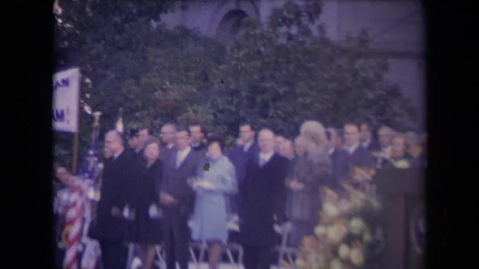 Home-movie frame of formally dressed attendees standing outdoors at a 1960s political ceremony, with an American flag and partial sign visible.