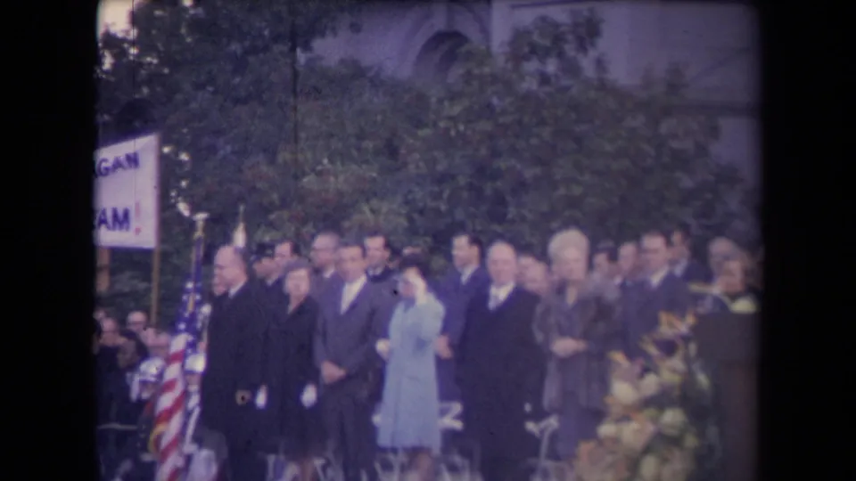 Formally dressed group stands near an American flag and partial sign during the Sacramento inauguration scene described by metadata.