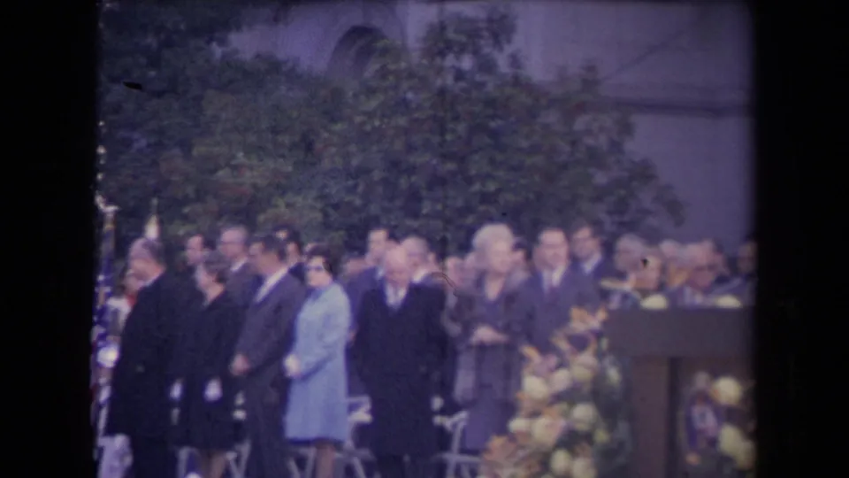 Soft 8mm frame showing a formal outdoor gathering with suited men, women in coats, folding chairs, flowers, and a podium area.