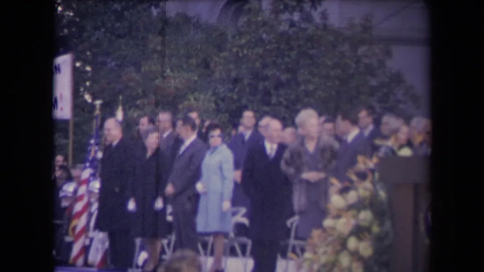 Outdoor 8mm ceremony frame with dignitaries, spectators, an American flag, partial sign, flowers, and podium in a Sacramento inauguration context.
