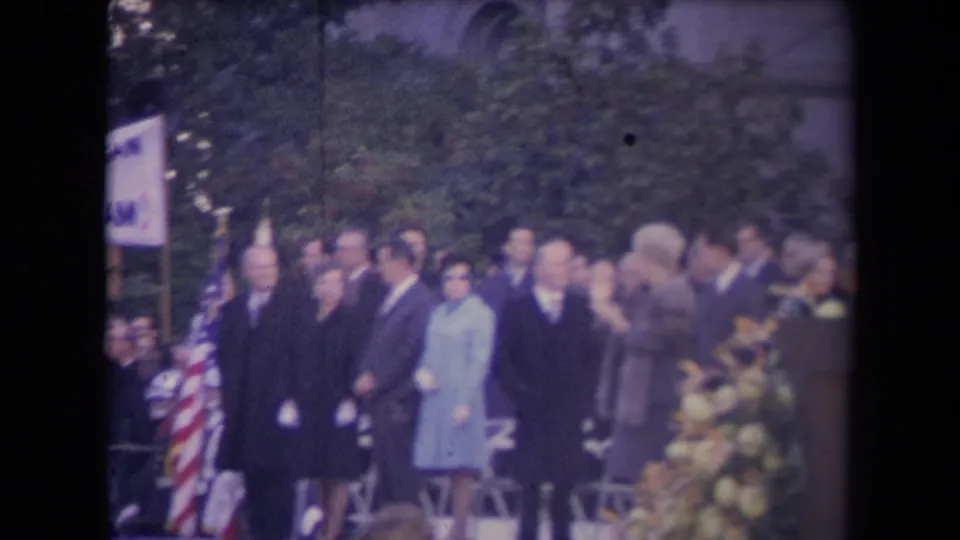 Soft 8mm frame of a formal outdoor ceremony with suited attendees, women in coats, American flags, and flowers.