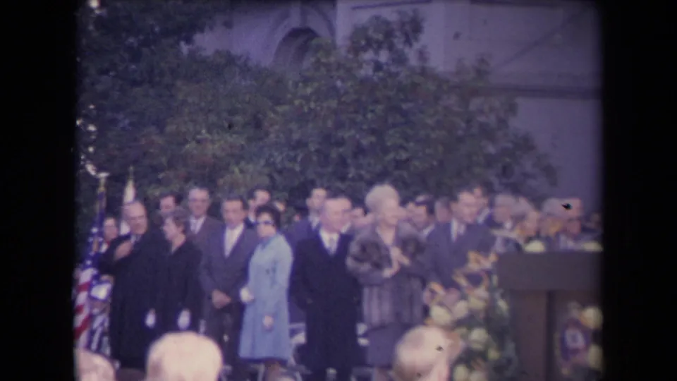 Formal crowd at a 1960s outdoor ceremony, with flags, flowers, a podium area, and a stone building behind them.