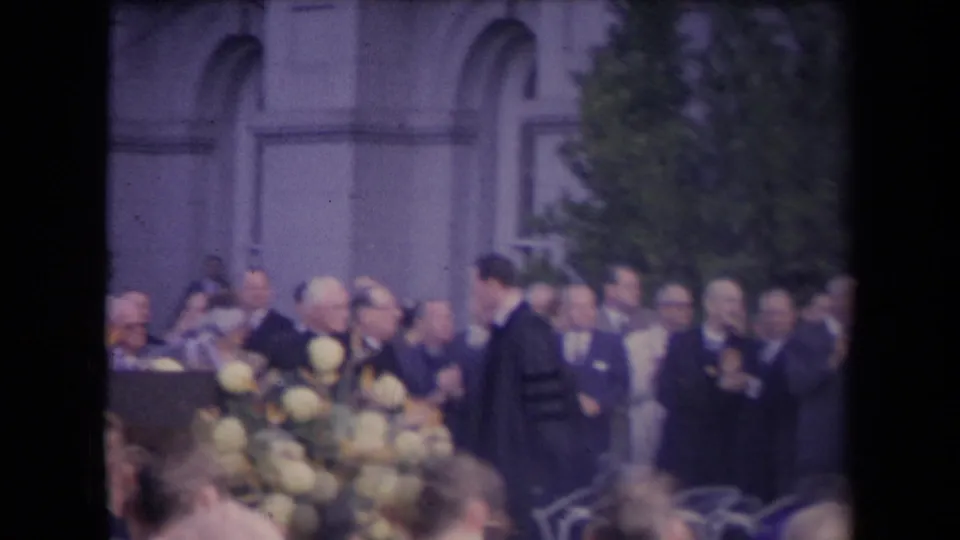 Handheld 8mm view of a formal outdoor audience applauding near flowers and arched stone architecture.